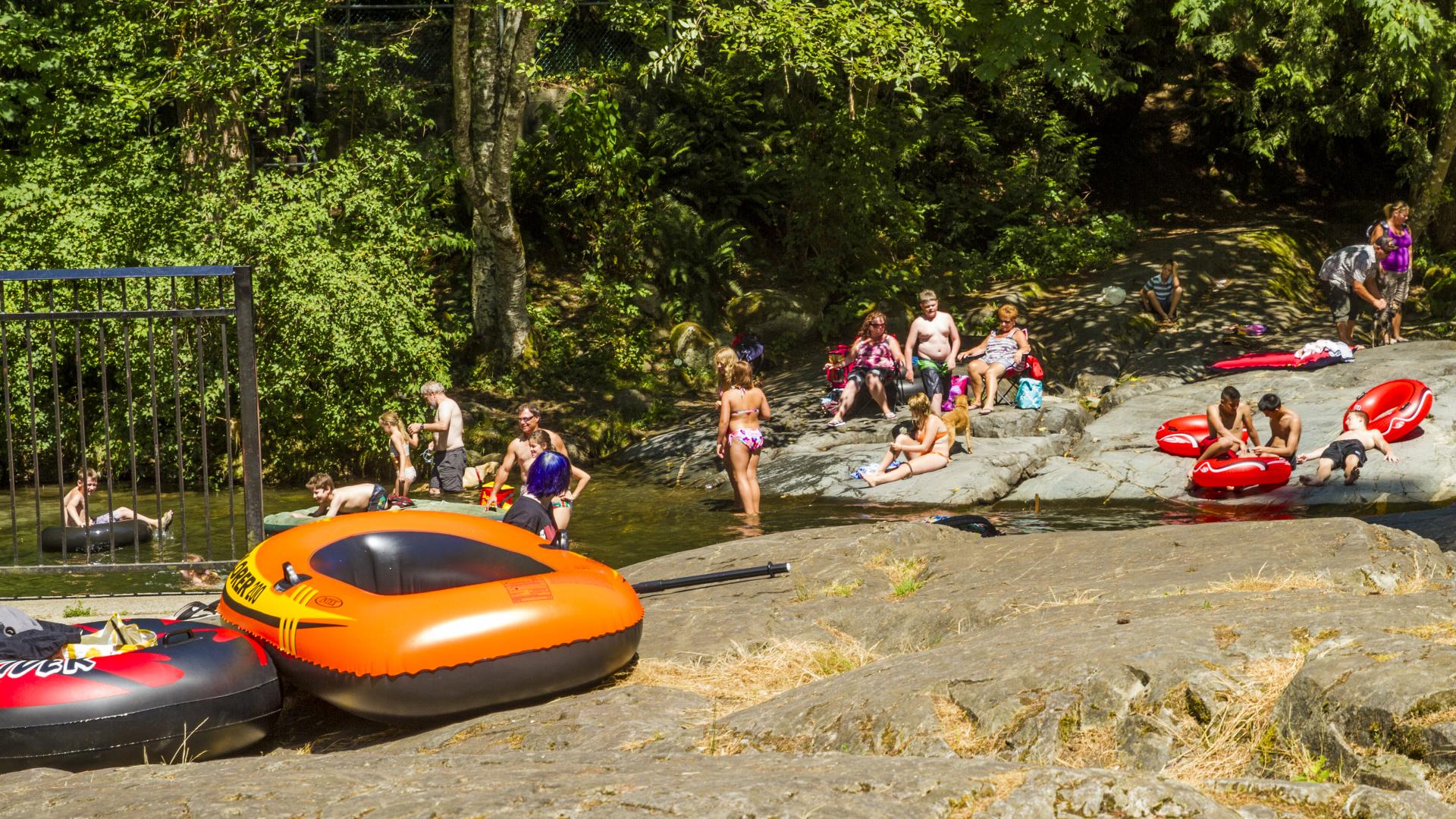 Several children, surrounded by large inflatable rafts stand and sit on the flat rocky shore of a river.